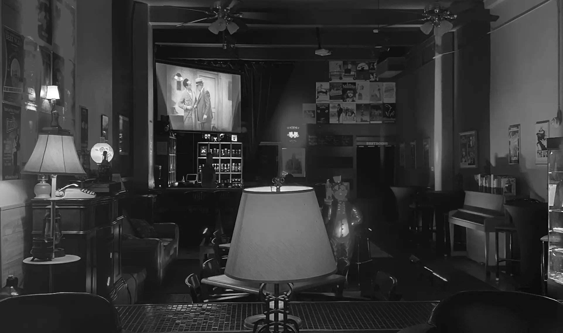 Matinee’s front room in low light — a vintage black-and-white film playing on the projection screen, a table lamp lit beside the armchairs, and framed film posters covering the walls