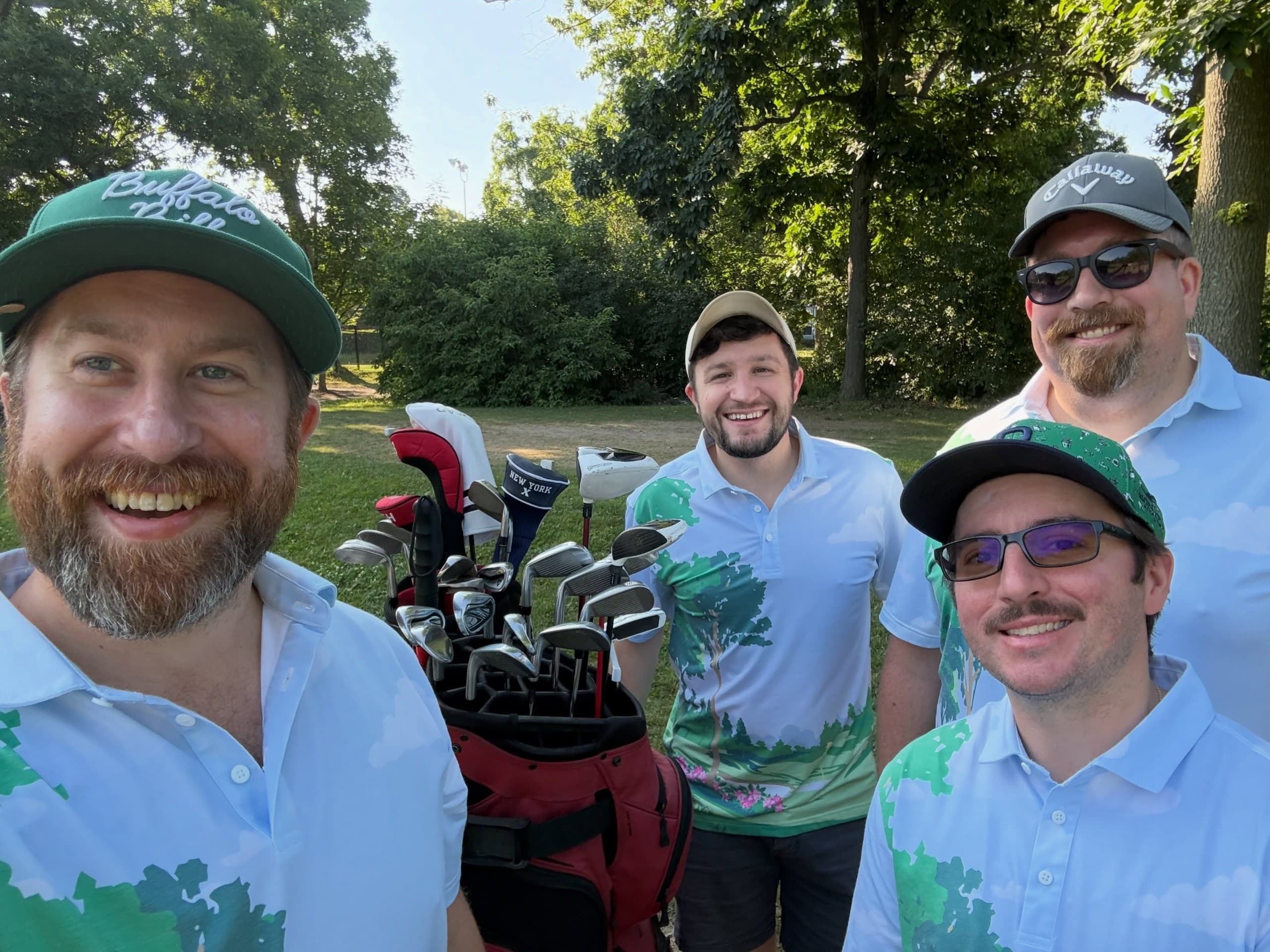 Five men wearing caps and light blue or green polo shirts smile at the camera at a golf course, with golf bags and clubs visible beside them.