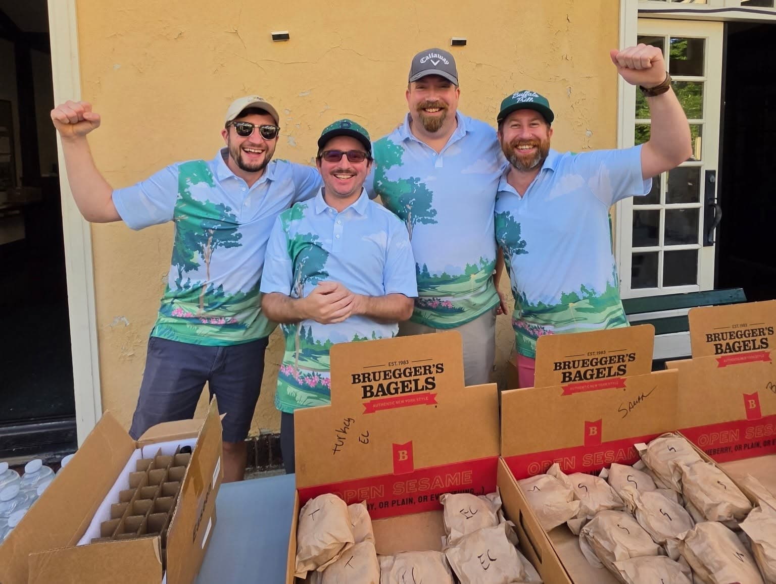 Four men wearing matching light blue shirts with green landscape designs and baseball caps stand with raised fists in front of a yellow building.