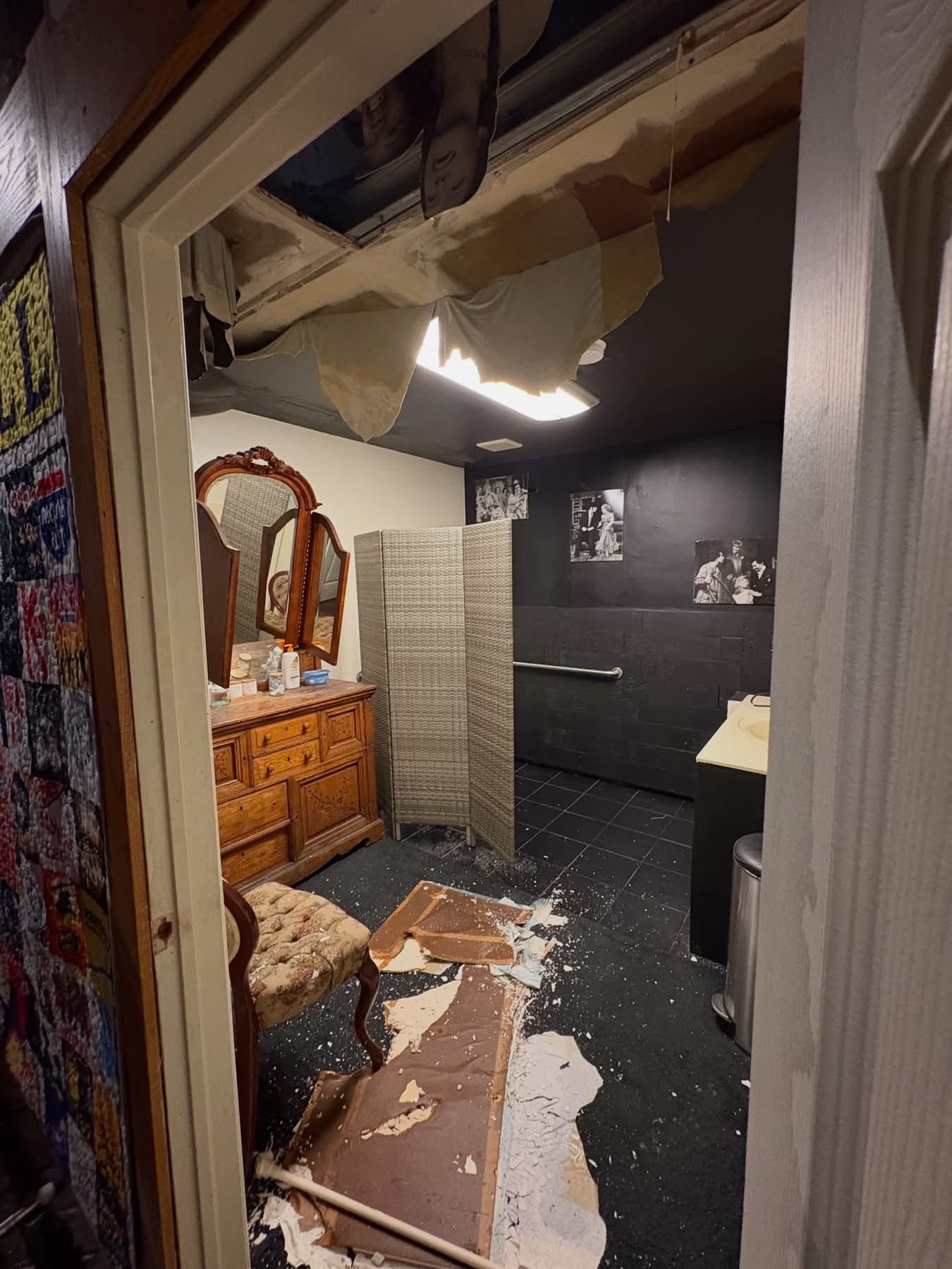 Bathroom undergoing renovation with damaged ceiling, exposed drywall, and debris on the floor. Wooden vanity with mirror, black tile walls, and shower area visible in background.