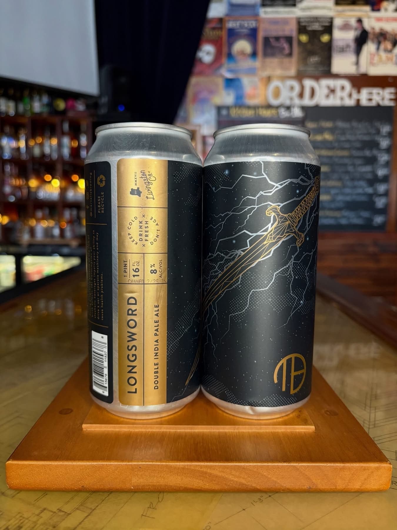 Two craft beer cans displayed on a wooden stand at a bar counter. The left can features gold and black labeling for Lonesword Double IPA.